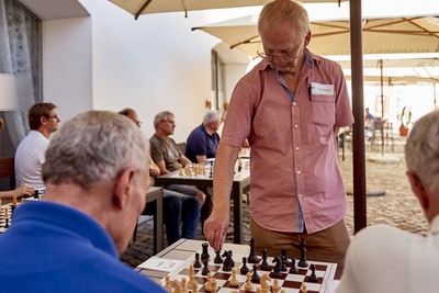 Großmeister mit Namensschild greift nach einer Figur an Brett 13, im Hintergrund weitere Spieler unter Schirmen