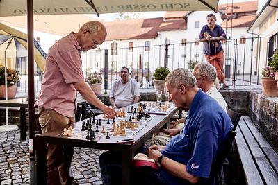 Großmeister zieht an einem Brett unter dem Sonnenschirm, ein Gegner im blauen Poloshirt grübelt, Zuschauer am Geländer