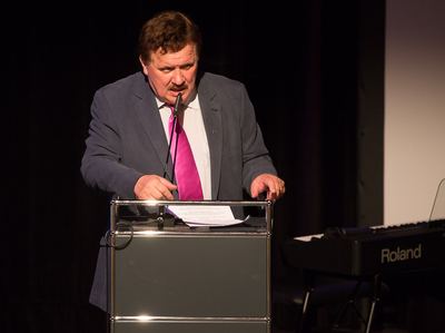 Redner mit pinkfarbener Krawatte spricht am Rednerpult, rechts im Bild ein Digitalpiano auf der Bühne