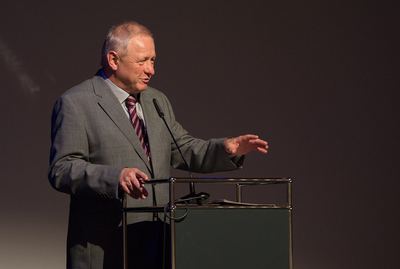 Redner im grauen Anzug mit gestreifter Krawatte spricht am Pult und gestikuliert mit einer Hand