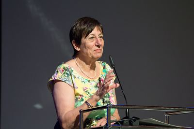 Rednerin im geblümten Kleid mit Perlenkette spricht am Mikrofon des Rednerpults