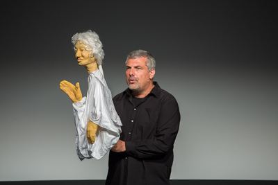 Der Sänger der Schachoper führt eine Handpuppe mit weißem Haar und weißem Gewand vor