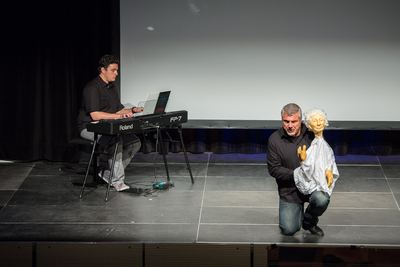 Der Sänger kniet mit einer weißhaarigen Handpuppe auf der Bühne, links begleitet der Pianist am Digitalpiano