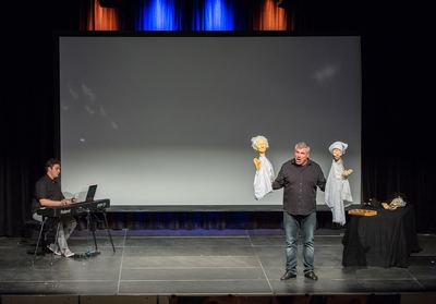 Bühne der Schachoper: Pianist am Digitalpiano, Sänger mit zwei weißen Puppen vor der Leinwand, rechts ein Tisch mit weiteren Figuren