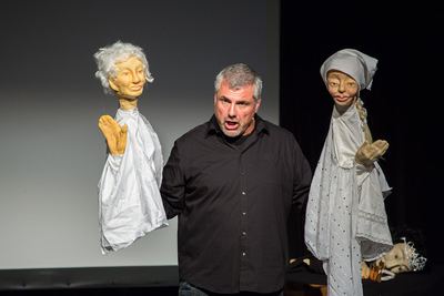 Sänger mit erhobenen Handpuppen, weißhaarige Figur und Bäuerin mit Kopftuch, hinten auf dem Tisch eine Puppe mit Krone