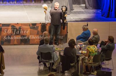 Sänger tritt mit zwei Puppen am Bühnenrand über dem Jubiläumsbanner auf, Ehrengäste am Tisch schauen beim Essen zu