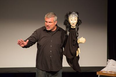 Sänger streckt abwehrend den Arm aus, an der anderen Hand die gekrönte Puppe mit schwarzem Haar und Gewand