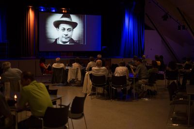 Gäste an Tischen sehen auf der Leinwand eine Schwarzweiß-Nahaufnahme eines Mannes mit Hut aus einem Stummfilm
