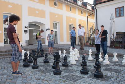 Gartenschach auf gepflastertem Hof vor einem gelb-weißen Gebäude, Spieler und Zuschauer stehen um die großen Figuren