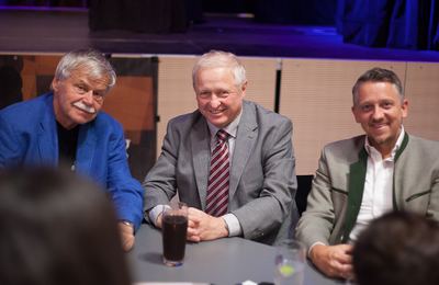 Drei Gäste sitzen lächelnd an einem Tisch vor der Bühne, vor ihnen ein Glas mit dunklem Getränk