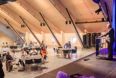 Redner am Pult spricht von der Bühne in den Saal mit hellem Holzdachstuhl, Gäste sitzen an verteilten Tischen