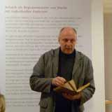 Redner mit aufgeschlagenem Buch vor einer Ausstellungswand mit Text zur Einführung in ein Schachgedicht aus dem 16. Jahrhundert